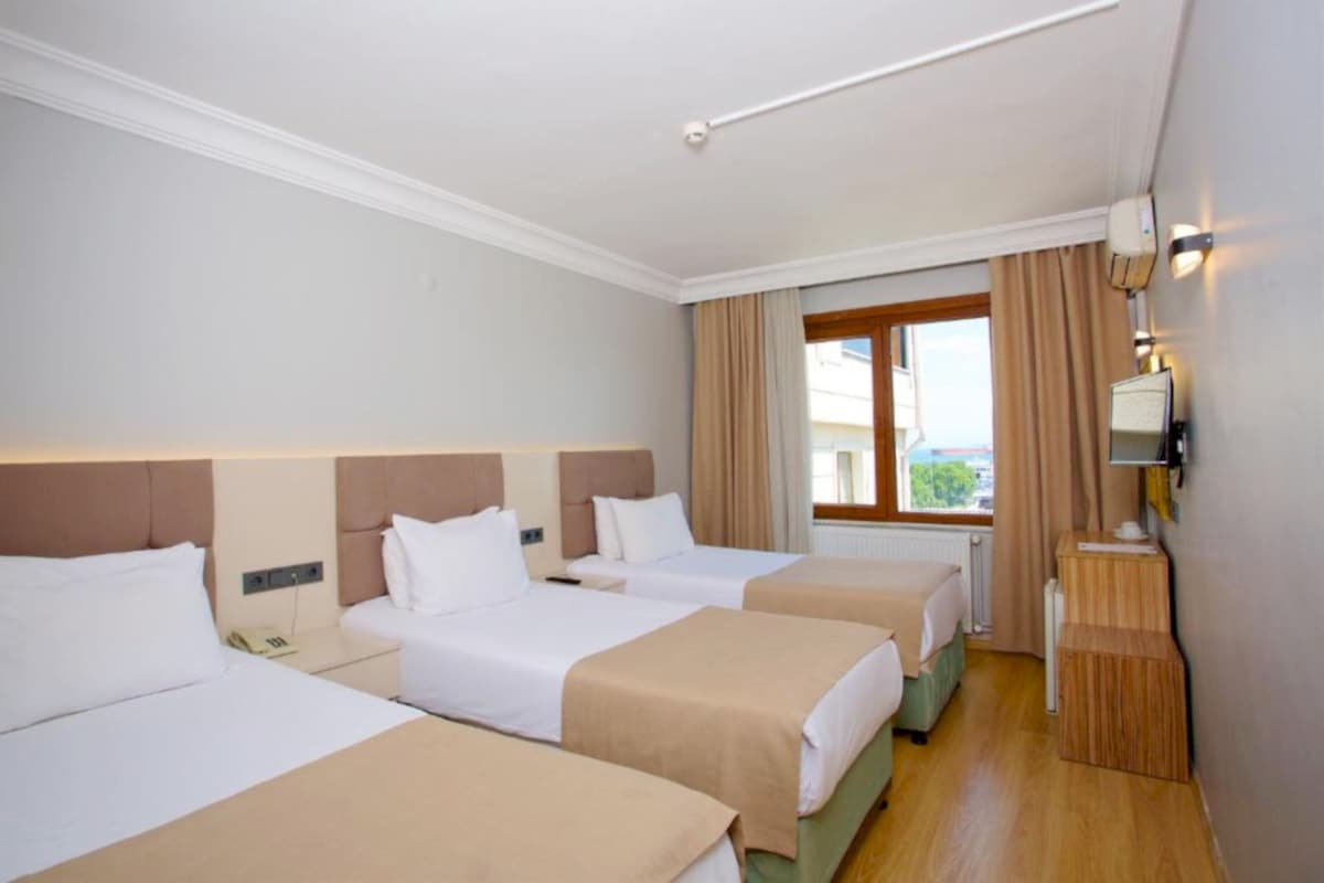 Una habitación de hotel con tres camas individuales, una mesita de noche de madera, una ventana con cortinas y un televisor montado en la pared.