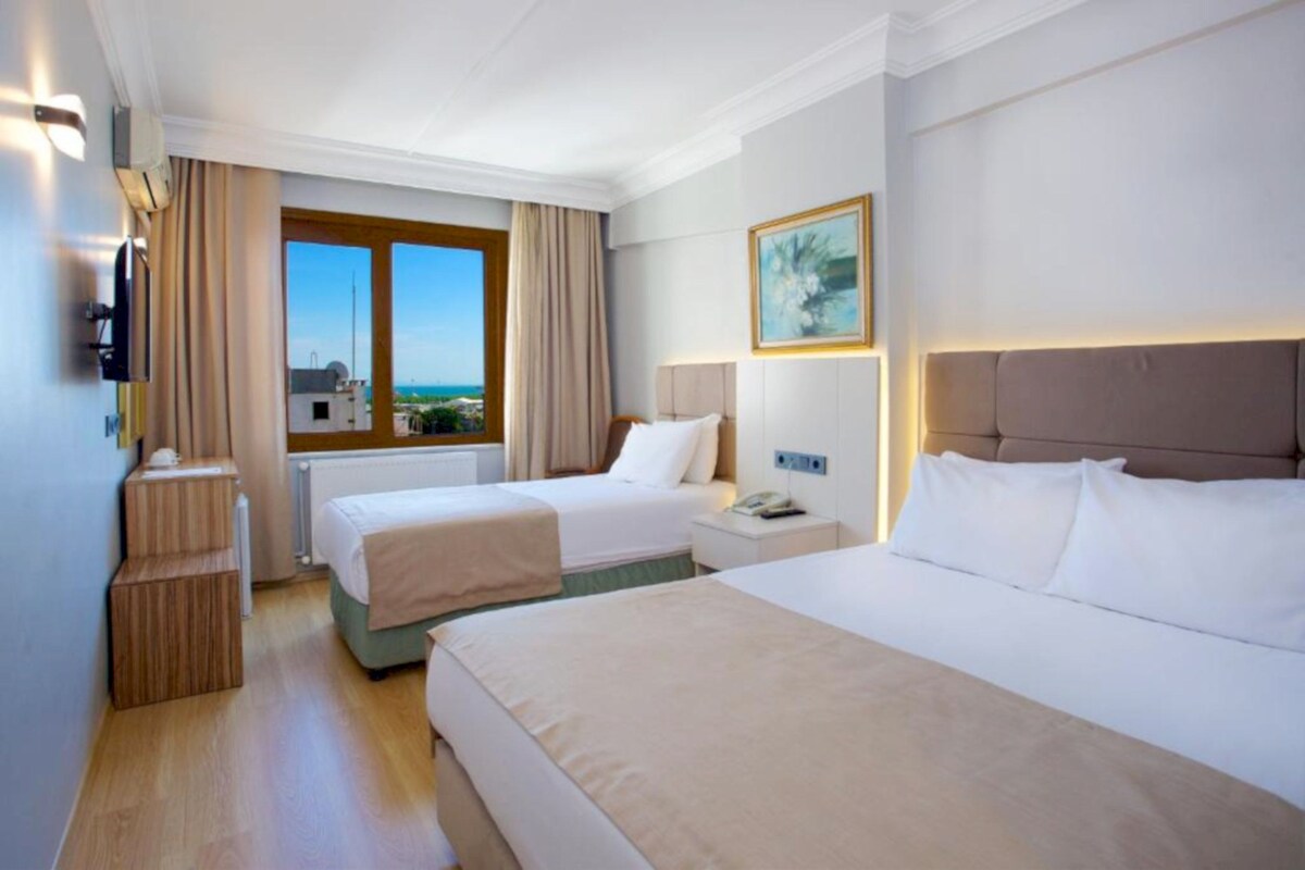 Habitación de hotel con dos camas, ventana con vistas, un cuadro en la pared y una mesita de noche con teléfono.