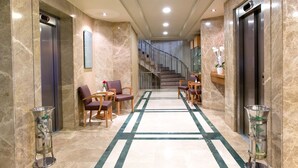 Un vestíbulo de hotel con piso de mármol, dos ascensores, una escalera y áreas de descanso con sillas y una mesa.