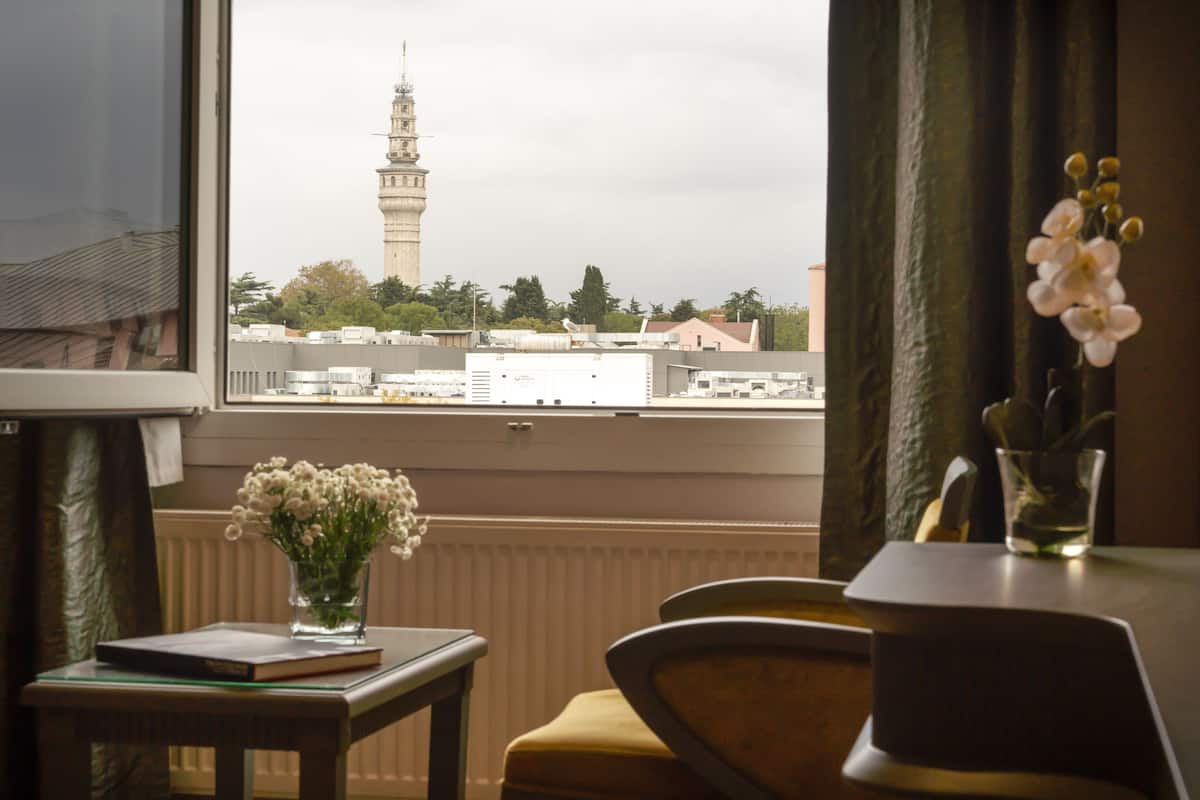 A room with a view of a tower through a window, a vase with flowers on a table, and a chair.