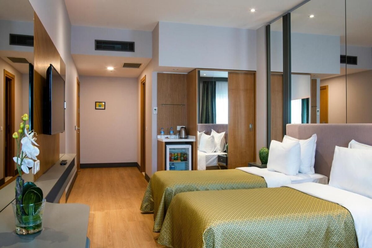 A hotel room with two beds, a bedside table, a mirror, a wardrobe, and a small kitchenette.