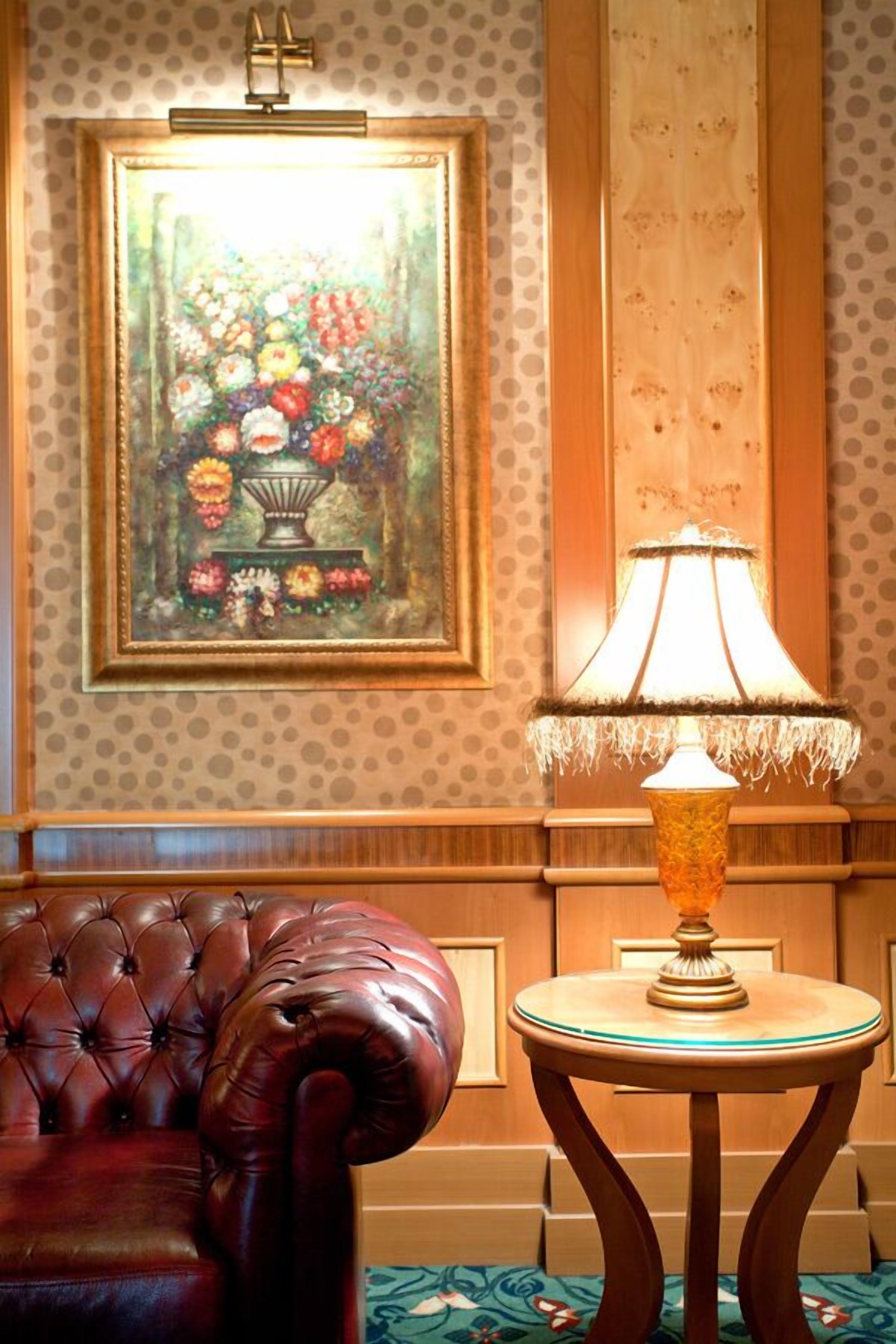 A room with a tufted leather armchair, a table with a lamp, and a framed floral painting on the wall.