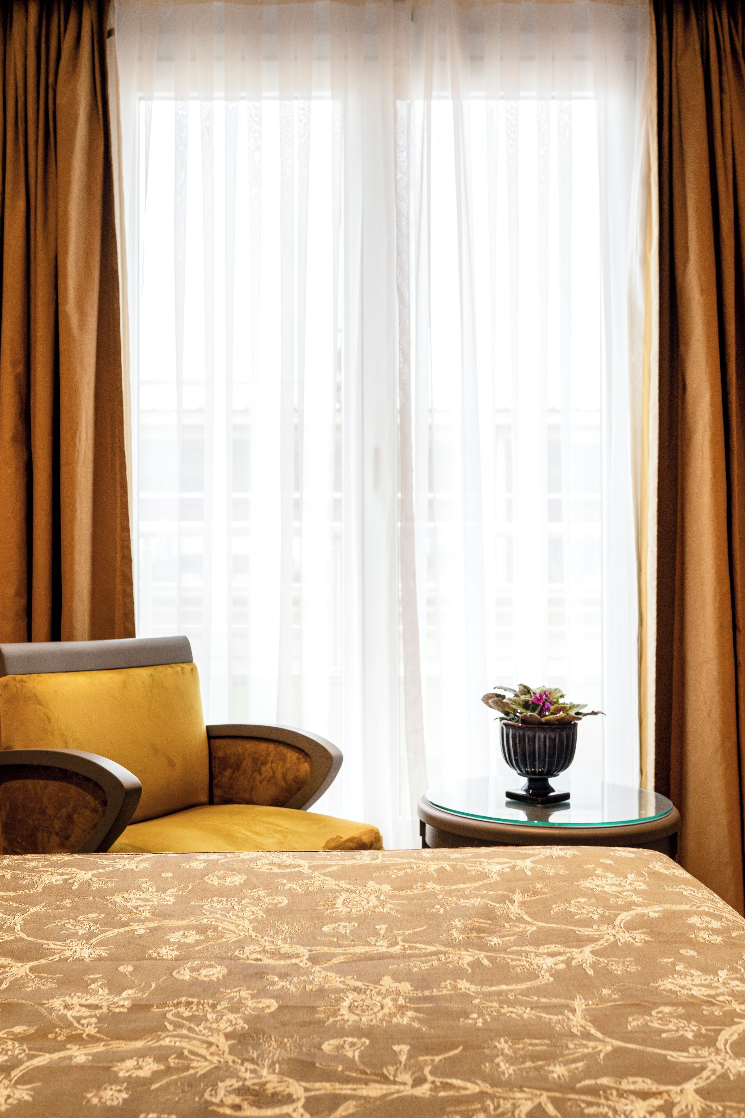 A hotel room with a bed, a chair, a small table with a plant, and sheer curtains.