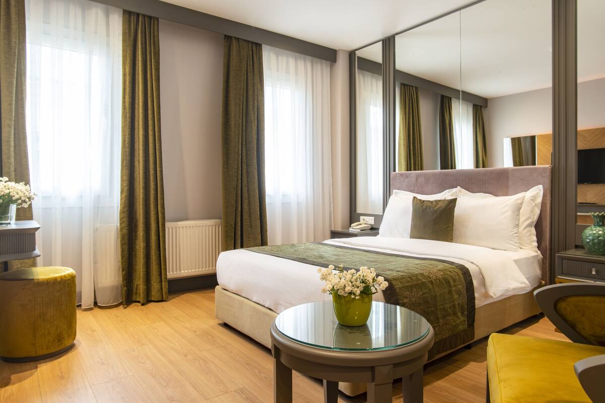 A hotel room with a large bed, a round glass table, a green vase with flowers, and a mirror on the wall.