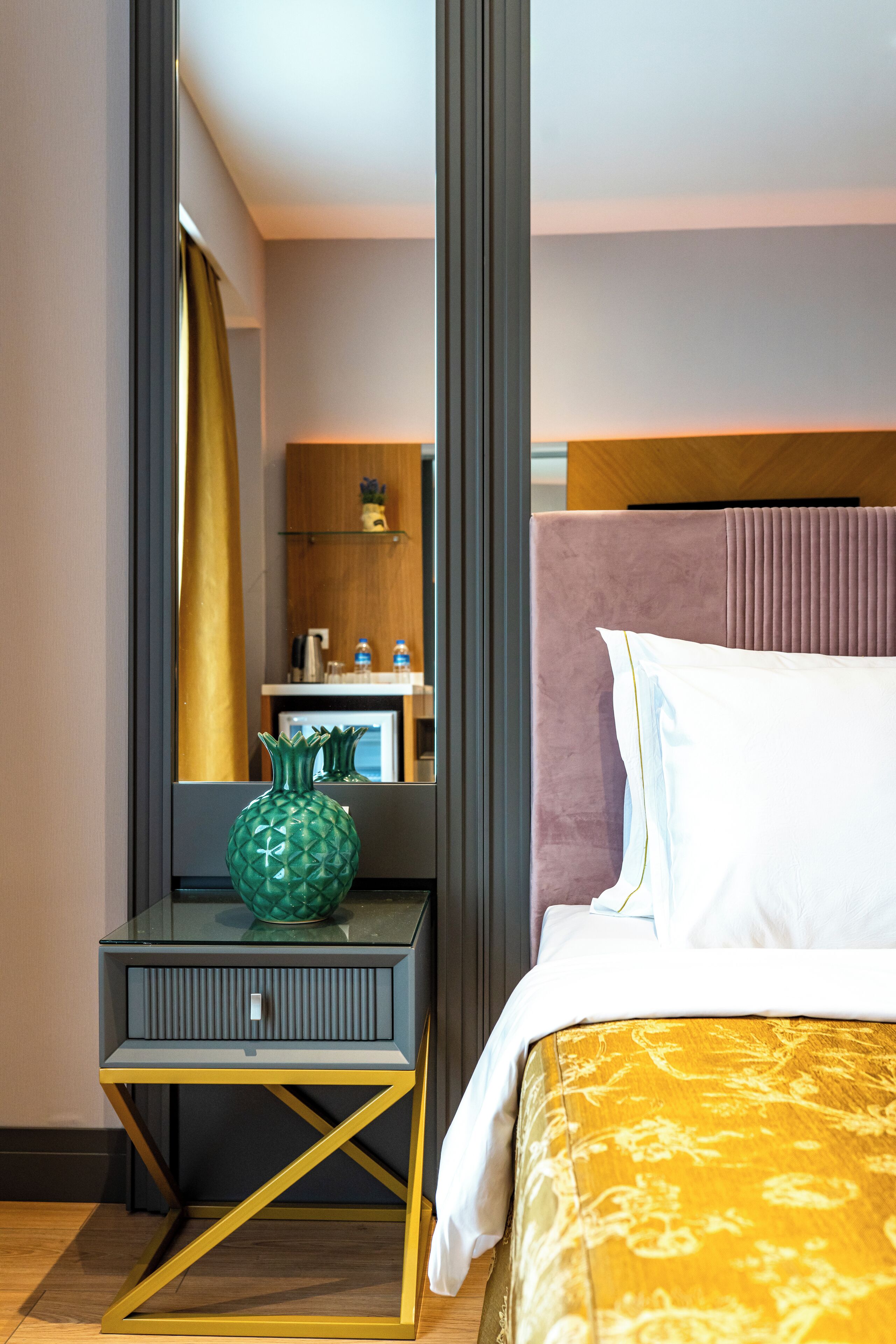 A hotel room with a bed, a nightstand, a mirror, and a small table with a green vase.