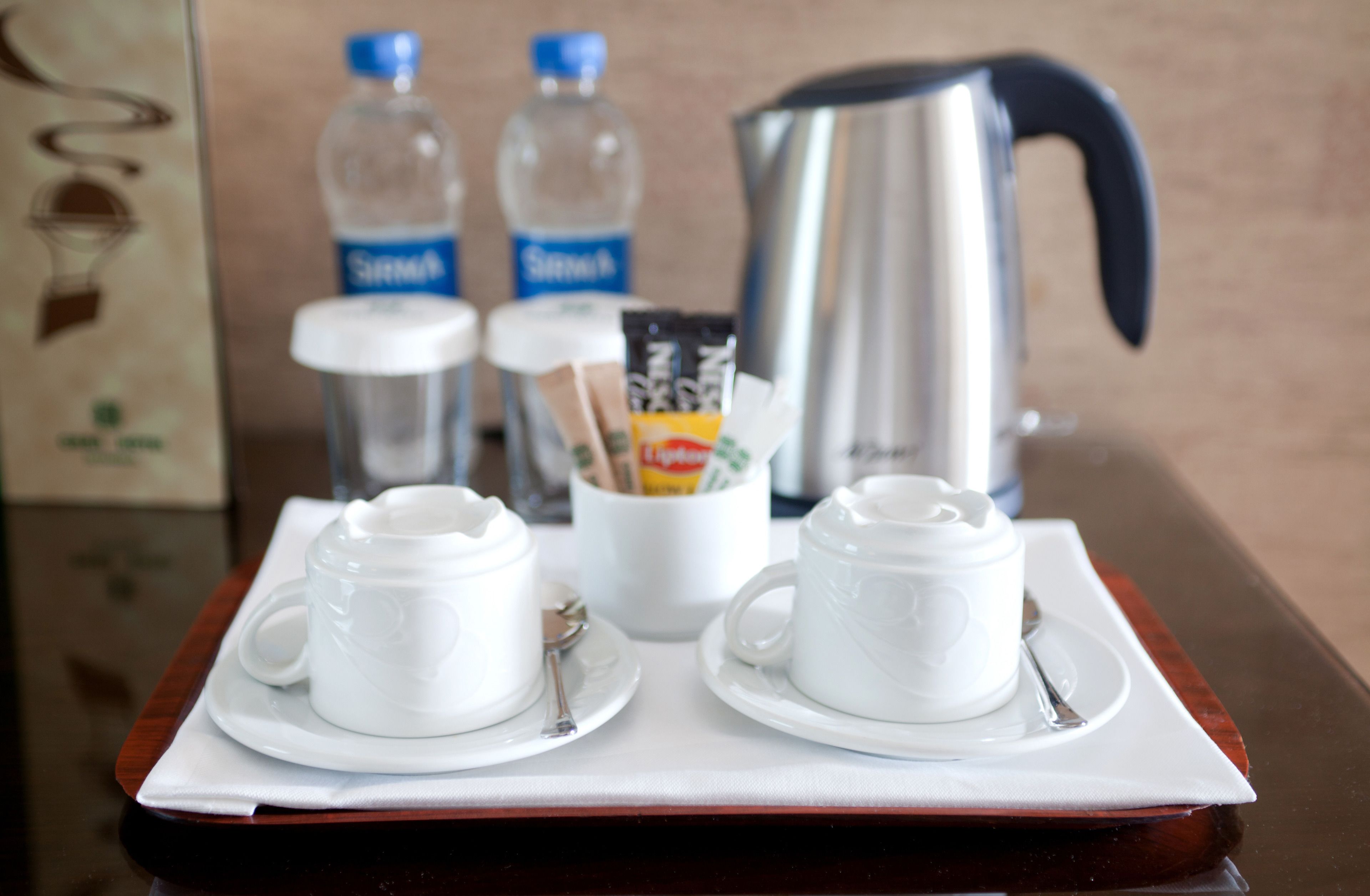 A tray with two white cups and saucers, a packet of sugar, two water bottles, and a silver electric kettle.