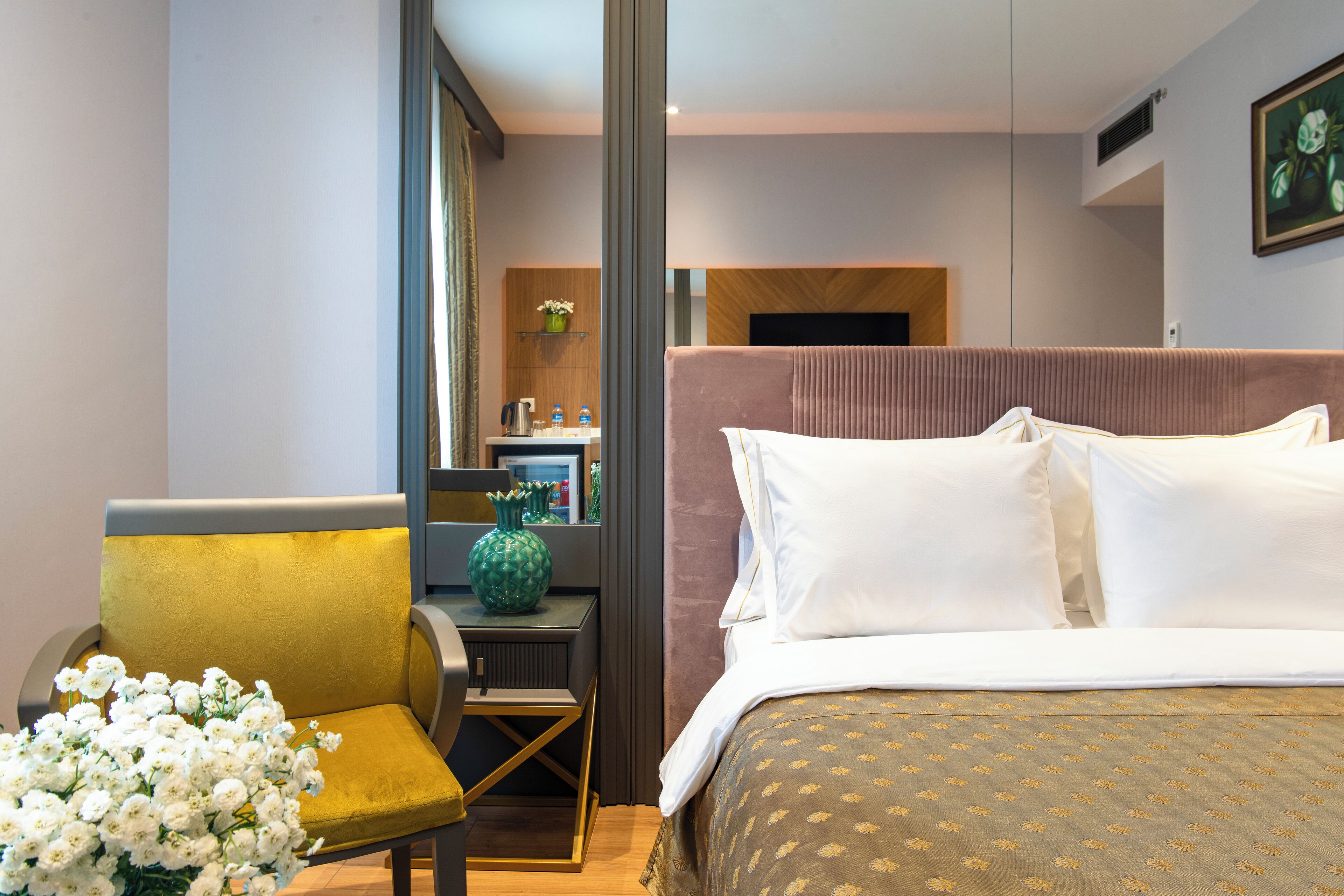 A hotel room with a bed, a chair, a small table, and a desk with a computer and a vase of flowers.
