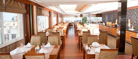 En restaurant i et hotel med flere borde og stole, et køkken i baggrunden og store vinduer.