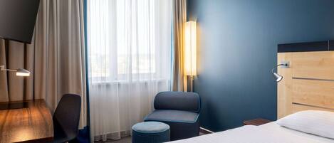 Une chambre d’hôtel comprenant un lit, un bureau, une chaise, un petit pouf, une lampe et une fenêtre avec des rideaux.