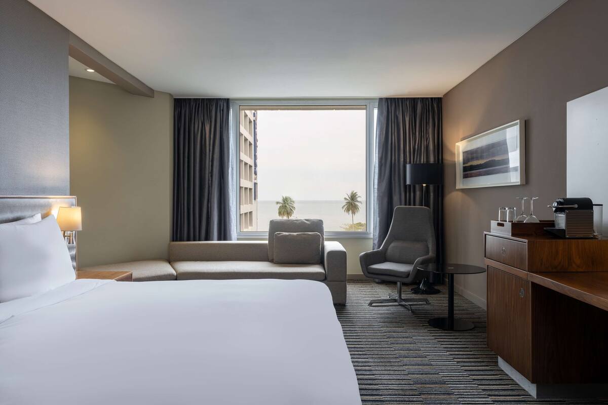 A hotel room with a large bed, a desk, a chair, a small table, a lamp, a view of the beach, and a window with curtains.