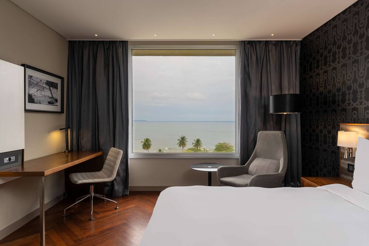 A hotel room with a large bed, two armchairs, a desk, a small table, and a view of the ocean.