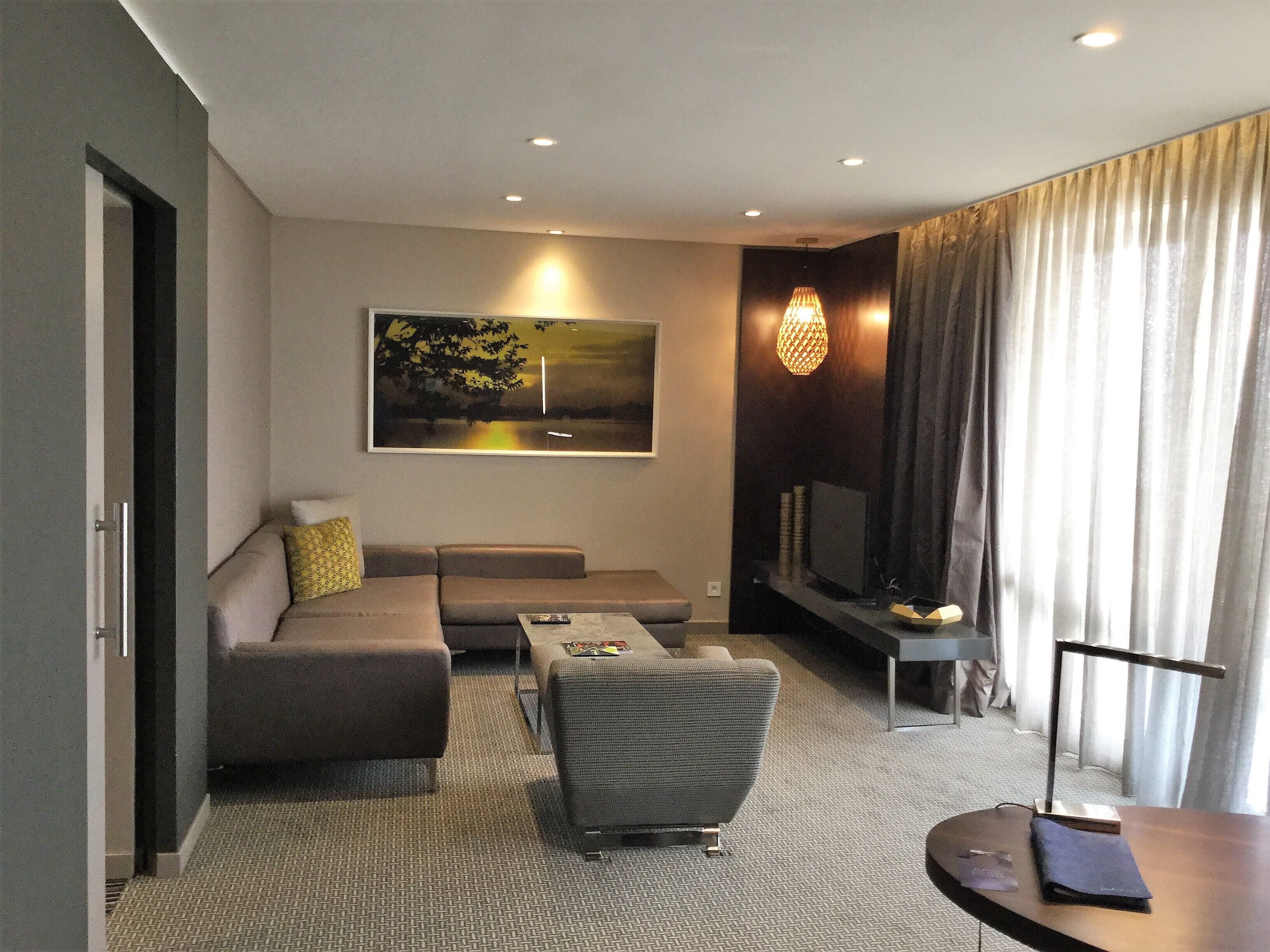 A modern living room with a sofa, armchair, and coffee table. A flat-screen TV is mounted on the wall, and there is a window with curtains.