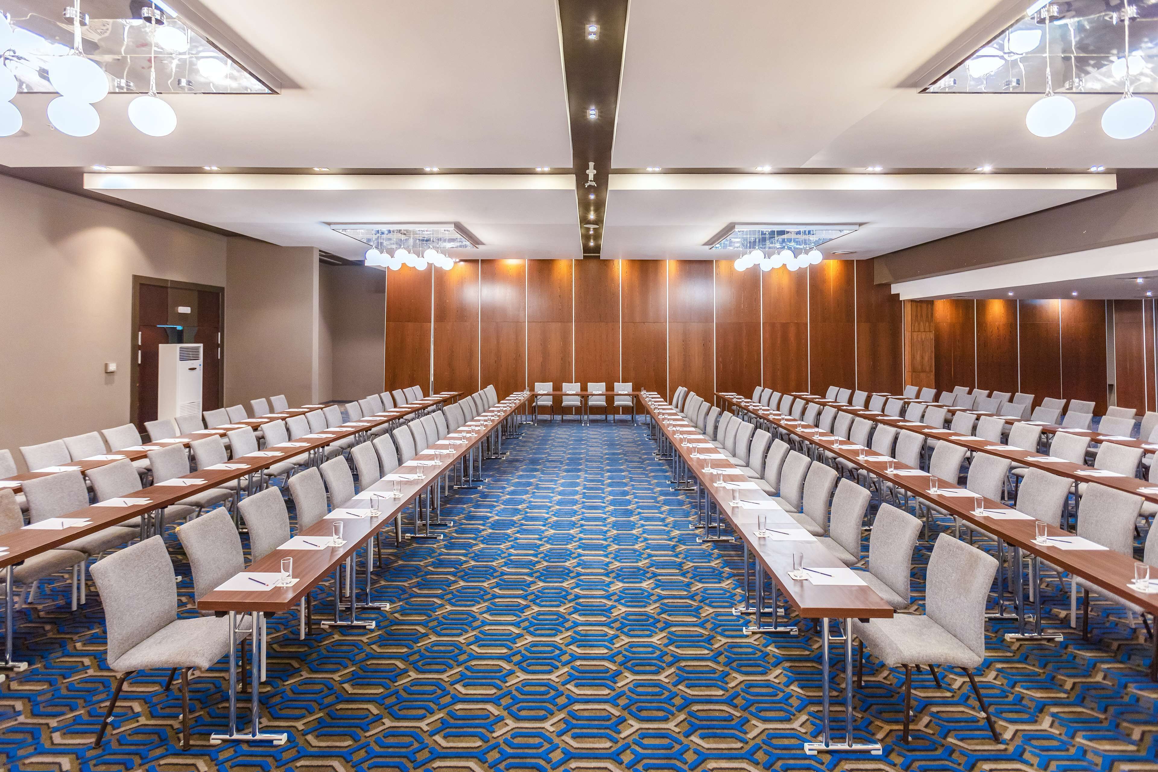 Ein Konferenzraum mit Tischen und Stühlen in Reihen, einem gemusterten Teppich und modernen Leuchten.