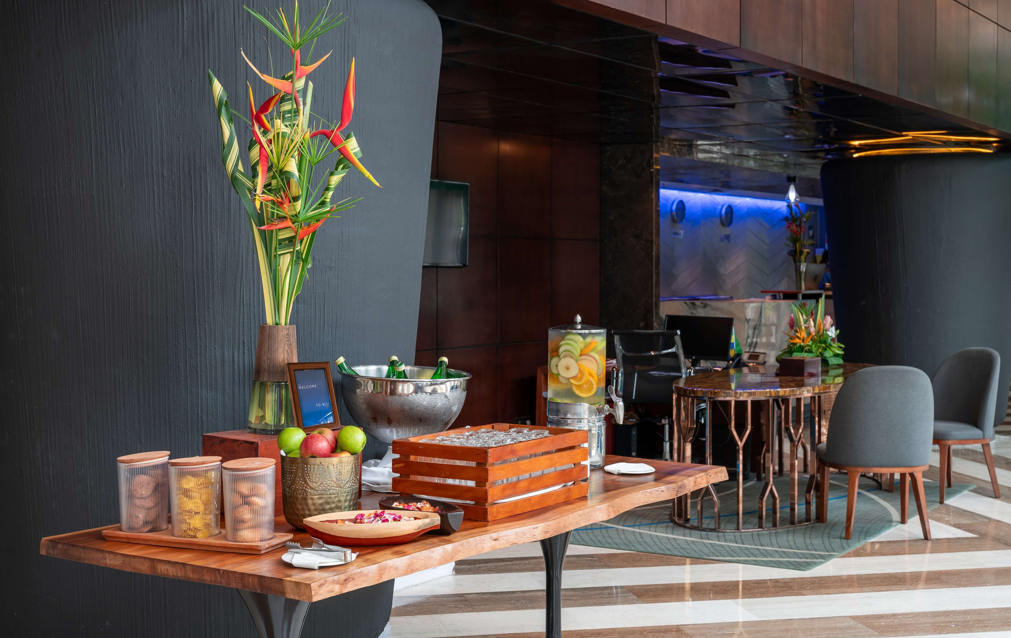 A reception area with a wooden table displaying a welcome sign, a decorative plant, a fruit bowl, and a beverage dispenser.