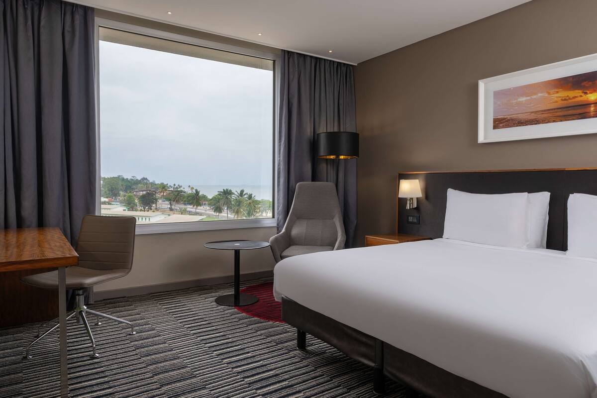 A hotel room with a large bed, two chairs, a small table, a lamp, and a view of the ocean.