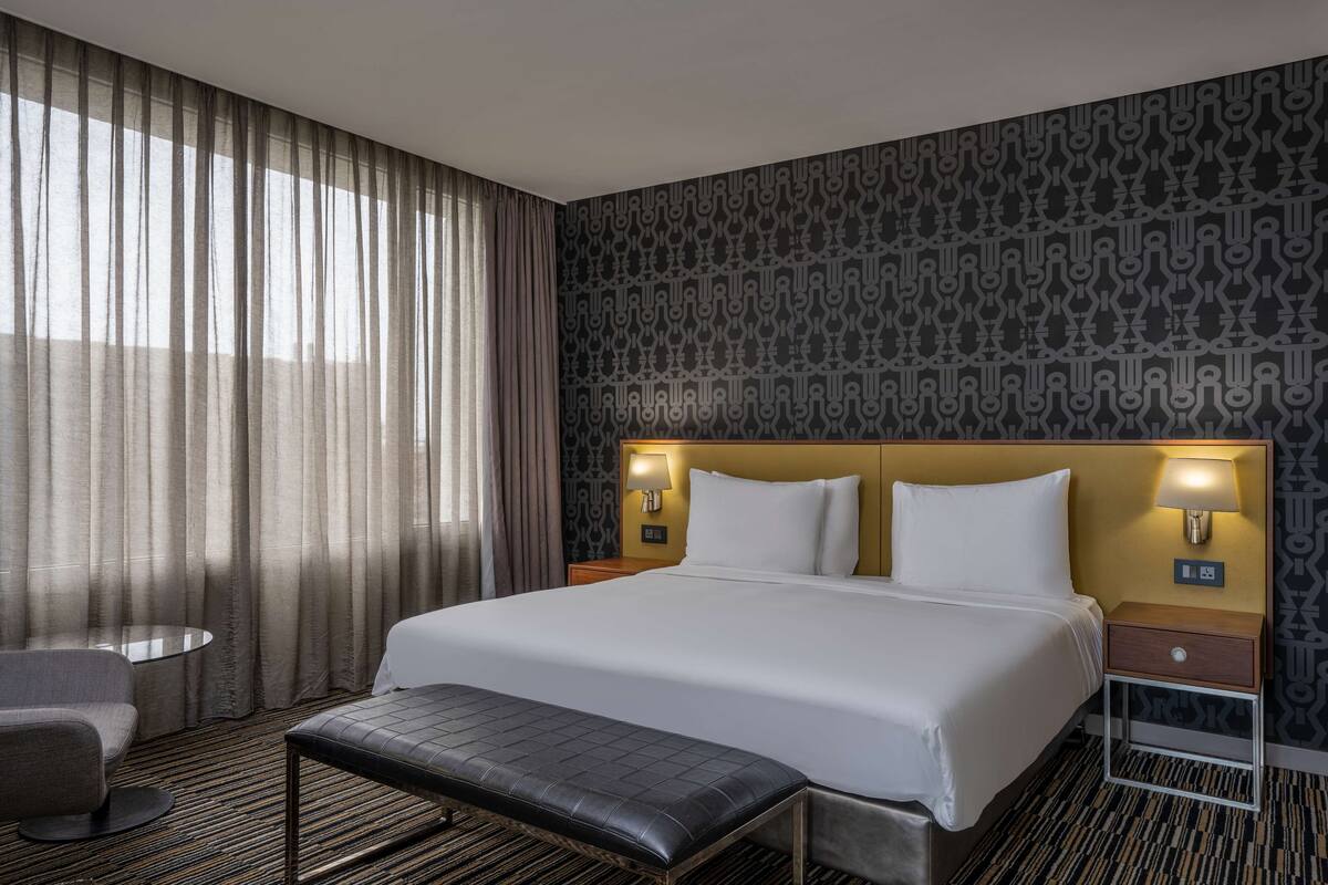 A hotel room with a large bed, two bedside lamps, a small round table, a bench, and a patterned wall.