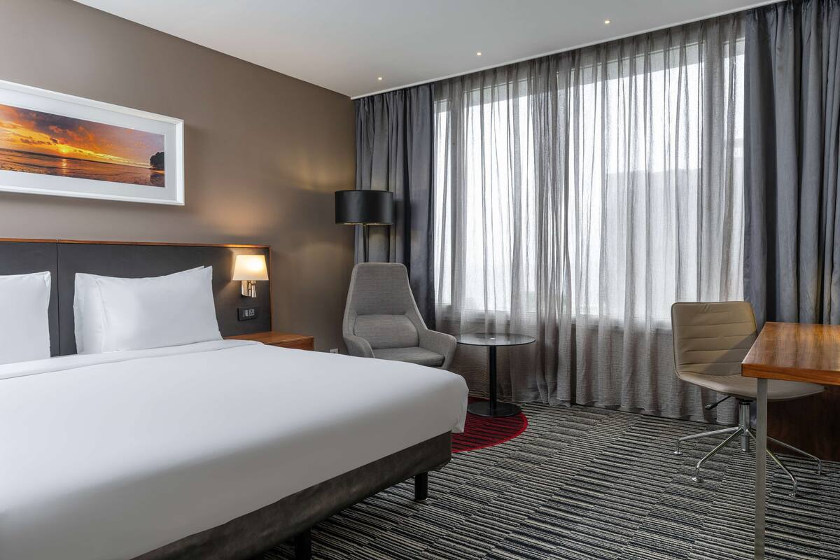 A hotel room with a large bed, two chairs, a small table, a lamp, and a painting of a sunset.
