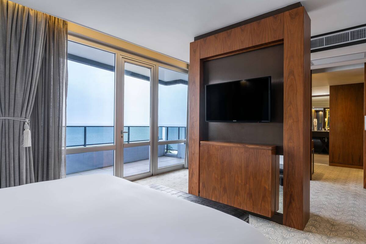 A hotel room with a large bed, a television mounted on a wooden panel, and a balcony with a view of the sea.