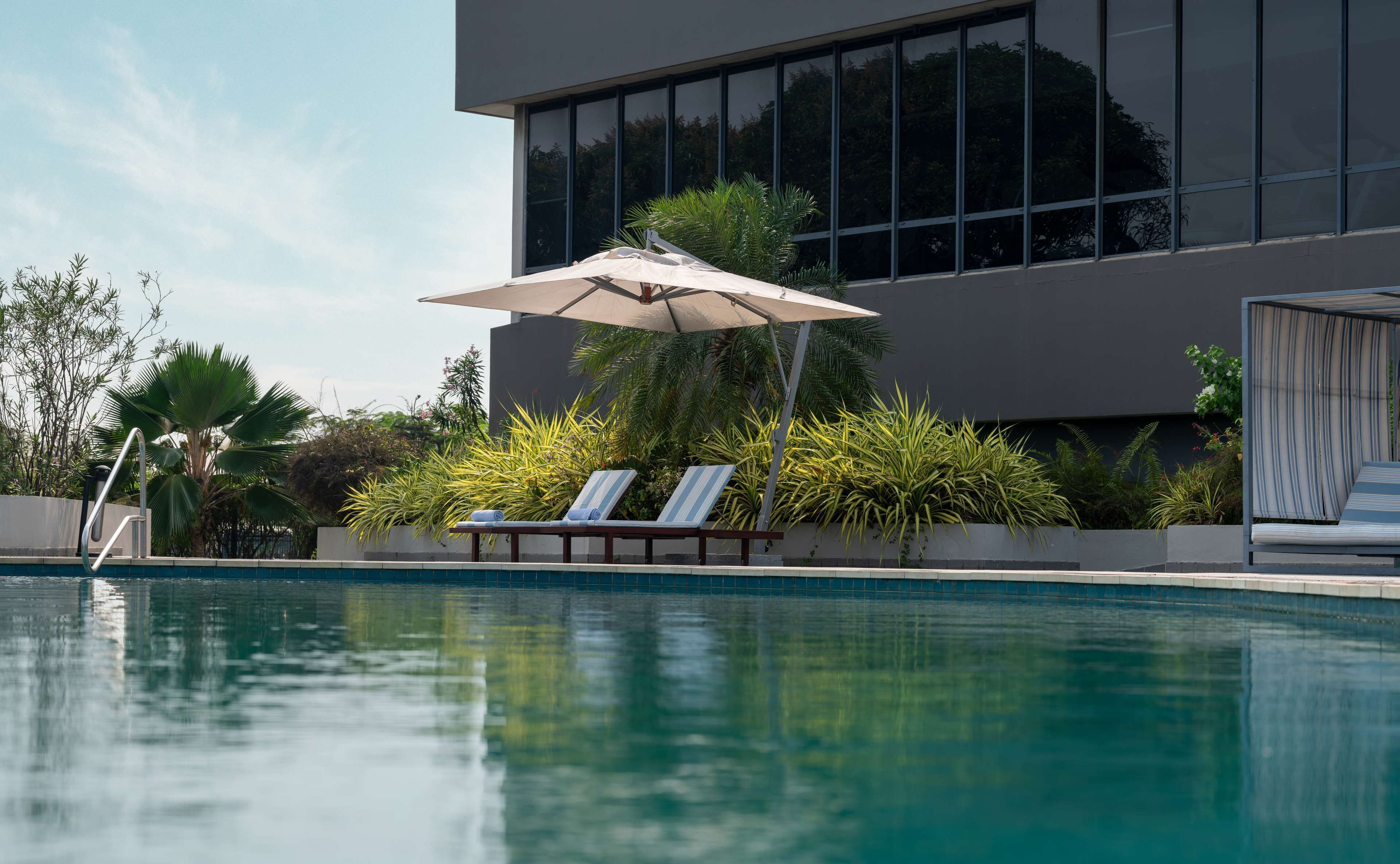 A modern pool area with two lounge chairs under a canopy, surrounded by lush greenery and a large building with reflective windows.