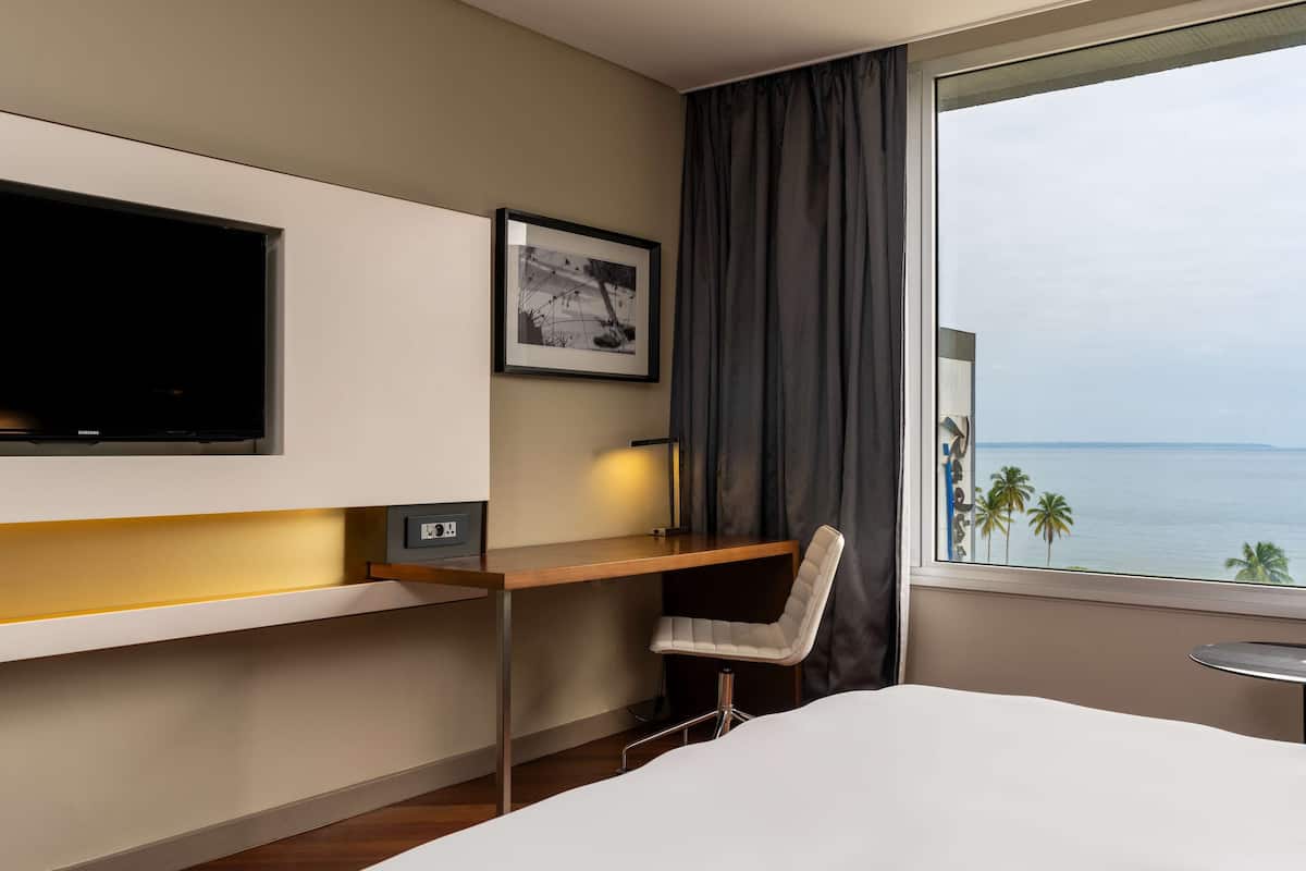 A hotel room with a bed, a desk with a chair, a TV, a framed picture, and a window with a view of the sea.