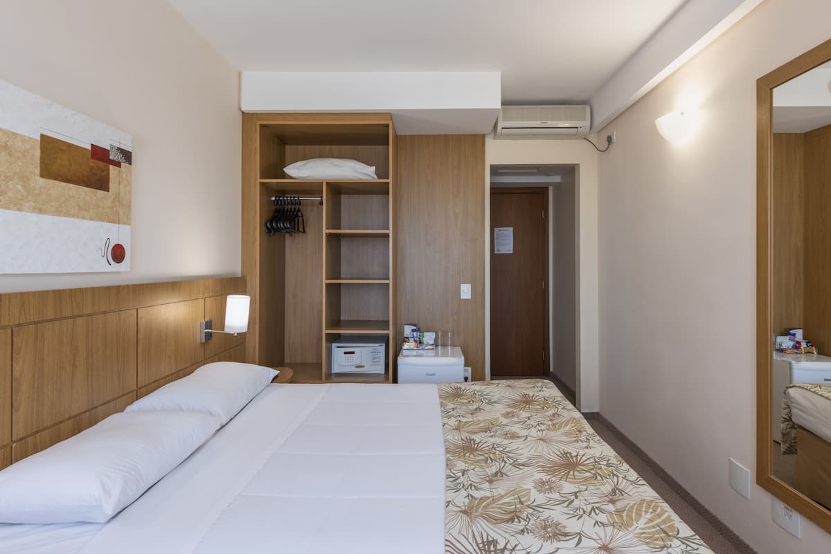 A hotel room with a bed, a wardrobe, a small desk with a lamp, and a mirror.
