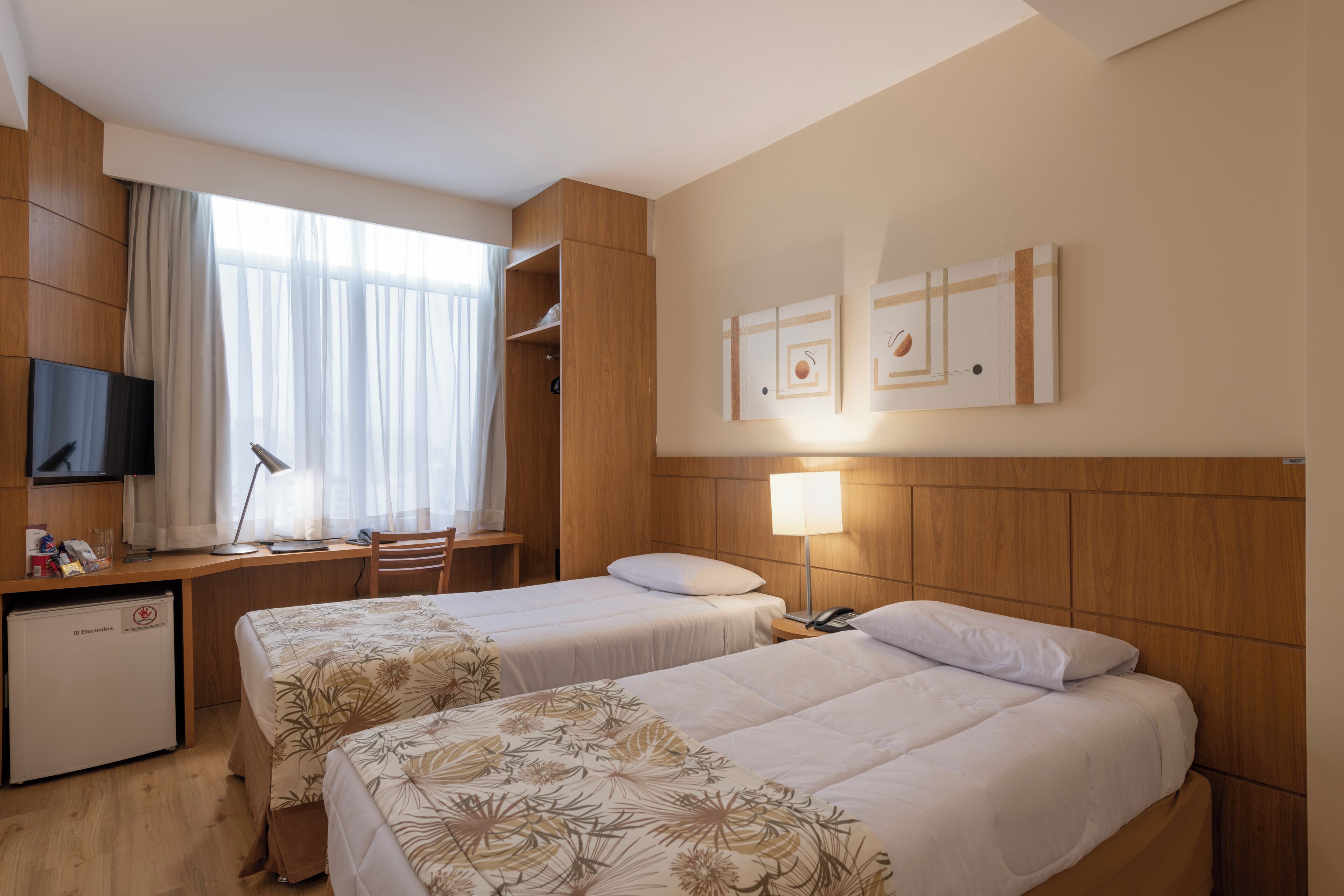 A hotel room with two beds, a desk, a television, and a small refrigerator.