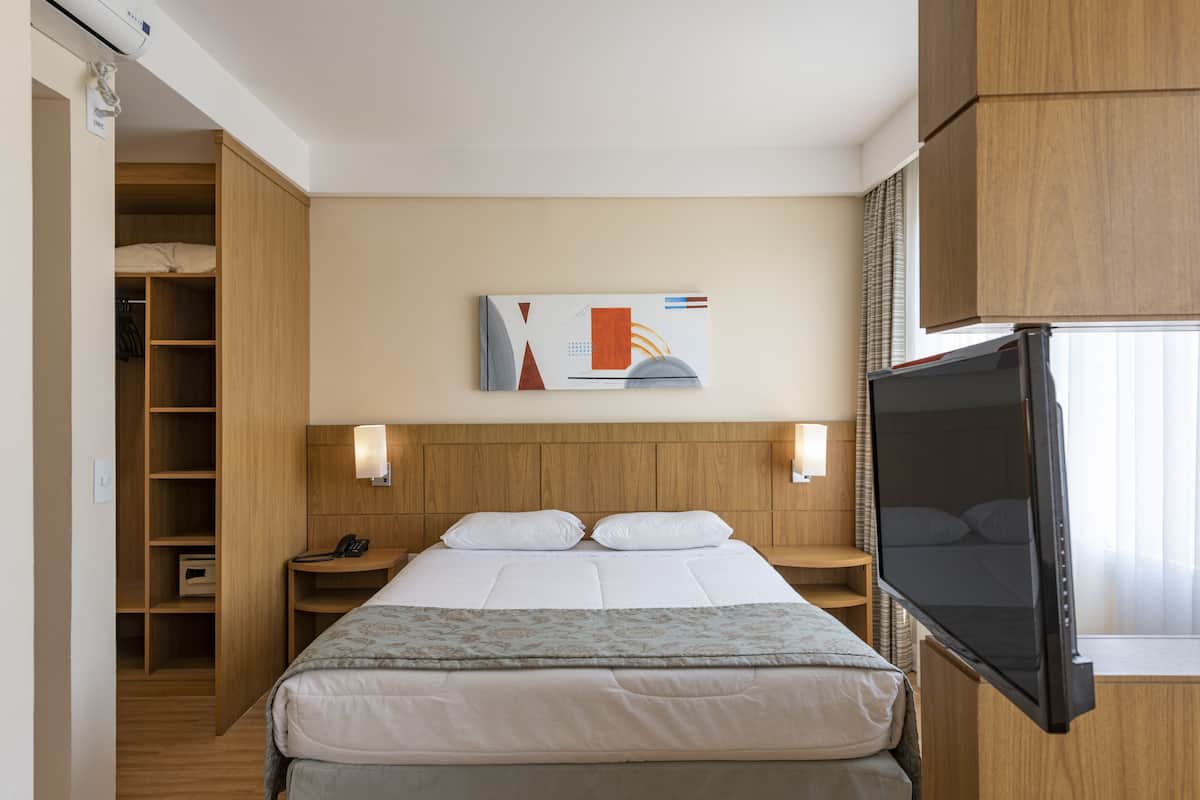 A hotel room with a bed, a TV, a wooden wardrobe, a bedside table, and a wall-mounted artwork.