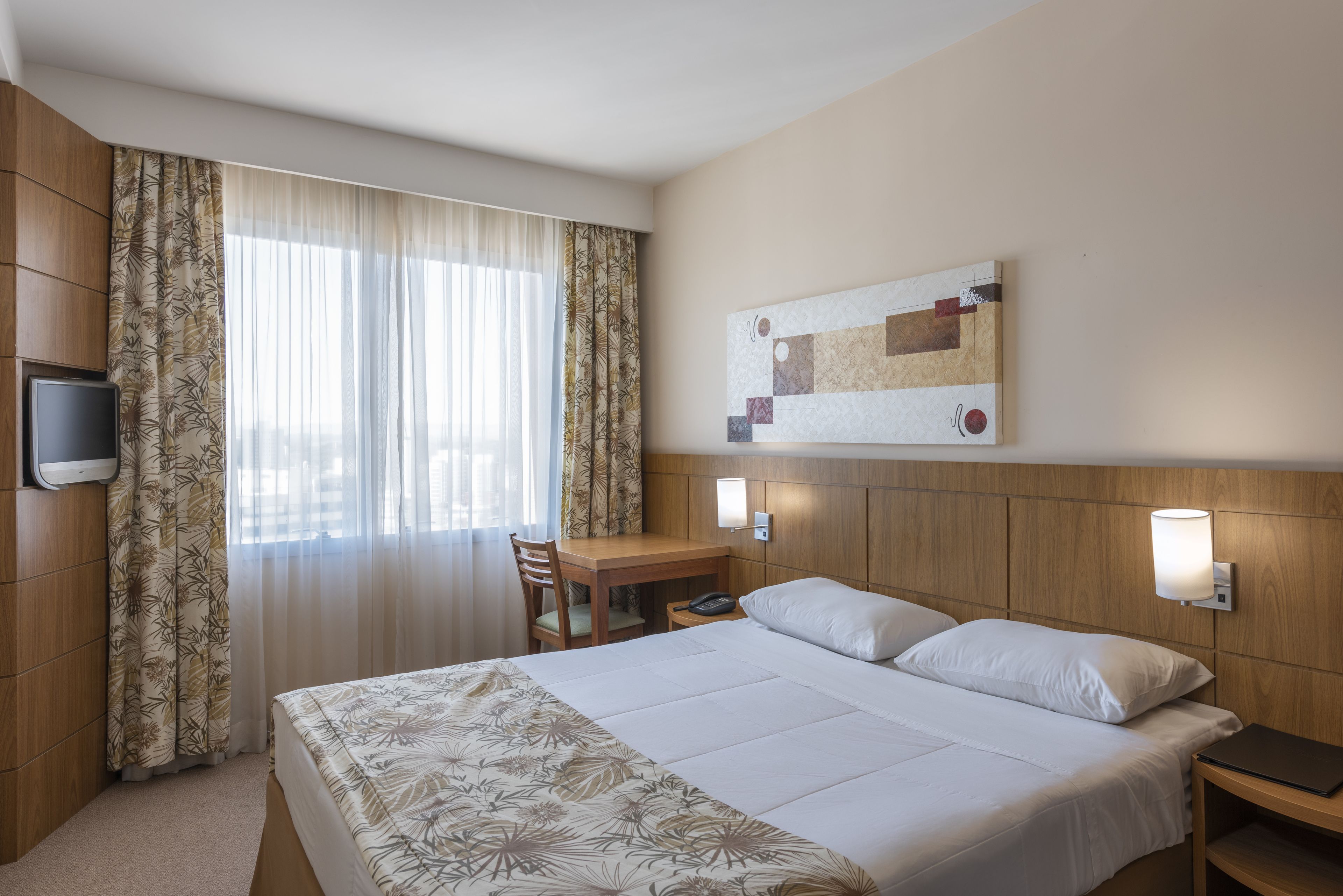 Habitación de hotel con cama, escritorio, silla, televisor y lámpara de pared.