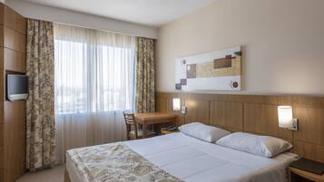 Habitación de hotel con cama, escritorio, silla, televisor y lámpara de pared.