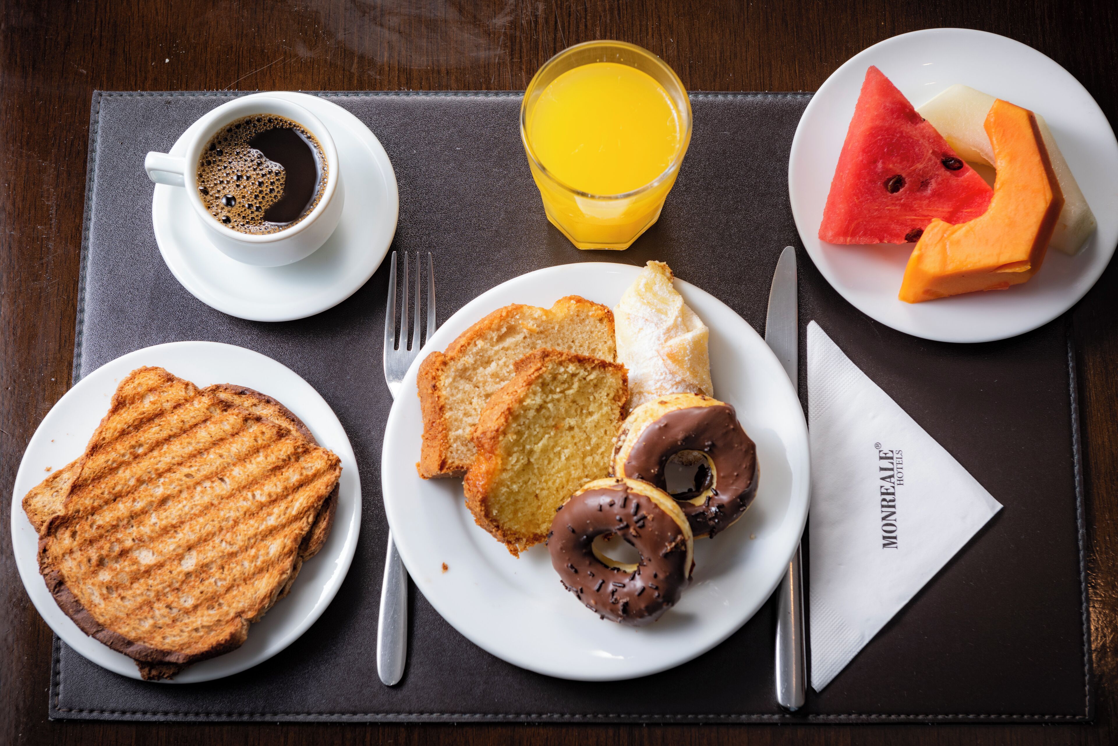 Une table dressée pour le petit-déjeuner avec des toasts, des œufs, des beignets, du café, du jus d’orange et des tranches de pastèque sur des assiettes.