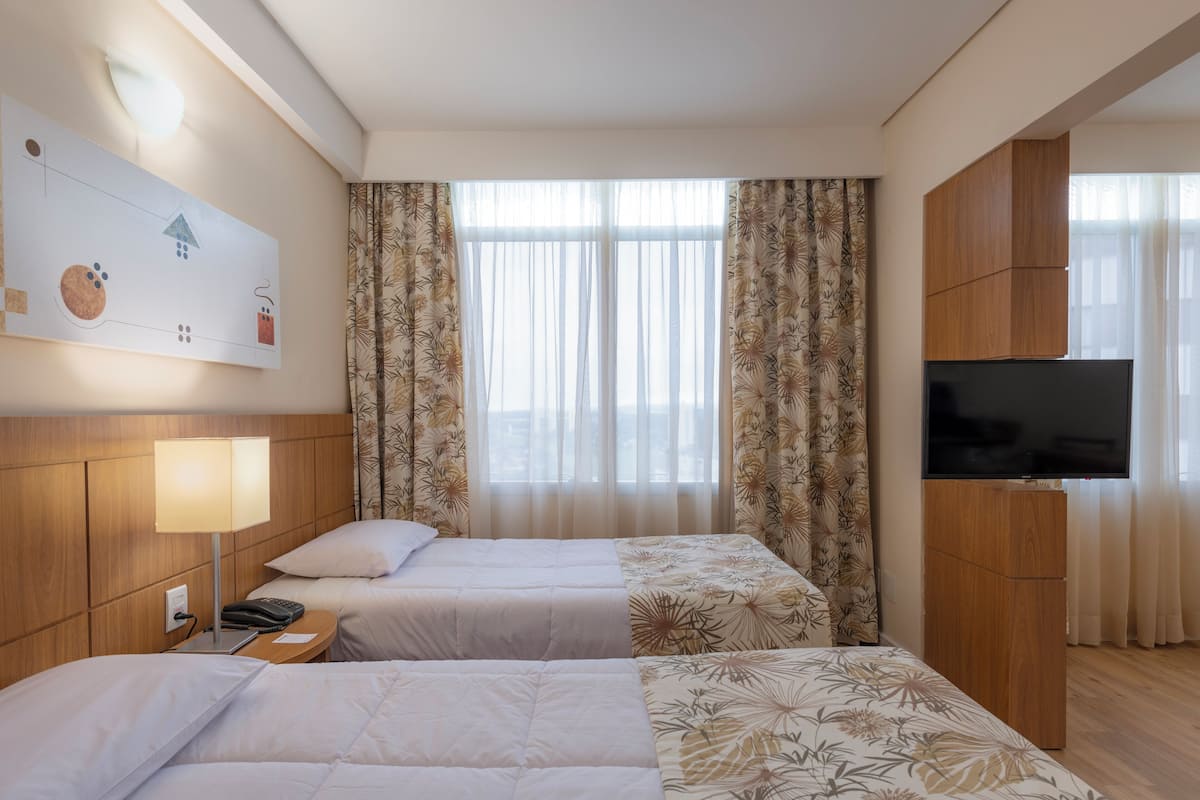 A hotel room with two beds, a TV, a desk with a lamp, and a wall-mounted artwork.