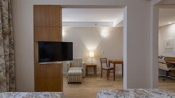 A compact hotel room with a flat-screen TV, a striped armchair, a small table with a lamp, and a wooden desk with a chair.