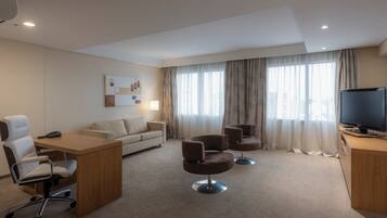 Una habitación de hotel moderna con un escritorio de madera, un sofá, dos sillones, un televisor y un amplio ventanal con cortinas.