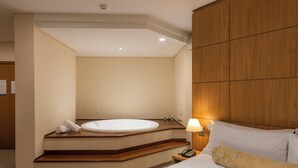 Habitación de hotel con una bañera grande, cabecero de madera, mesita de noche con lámpara y cama bien hecha.