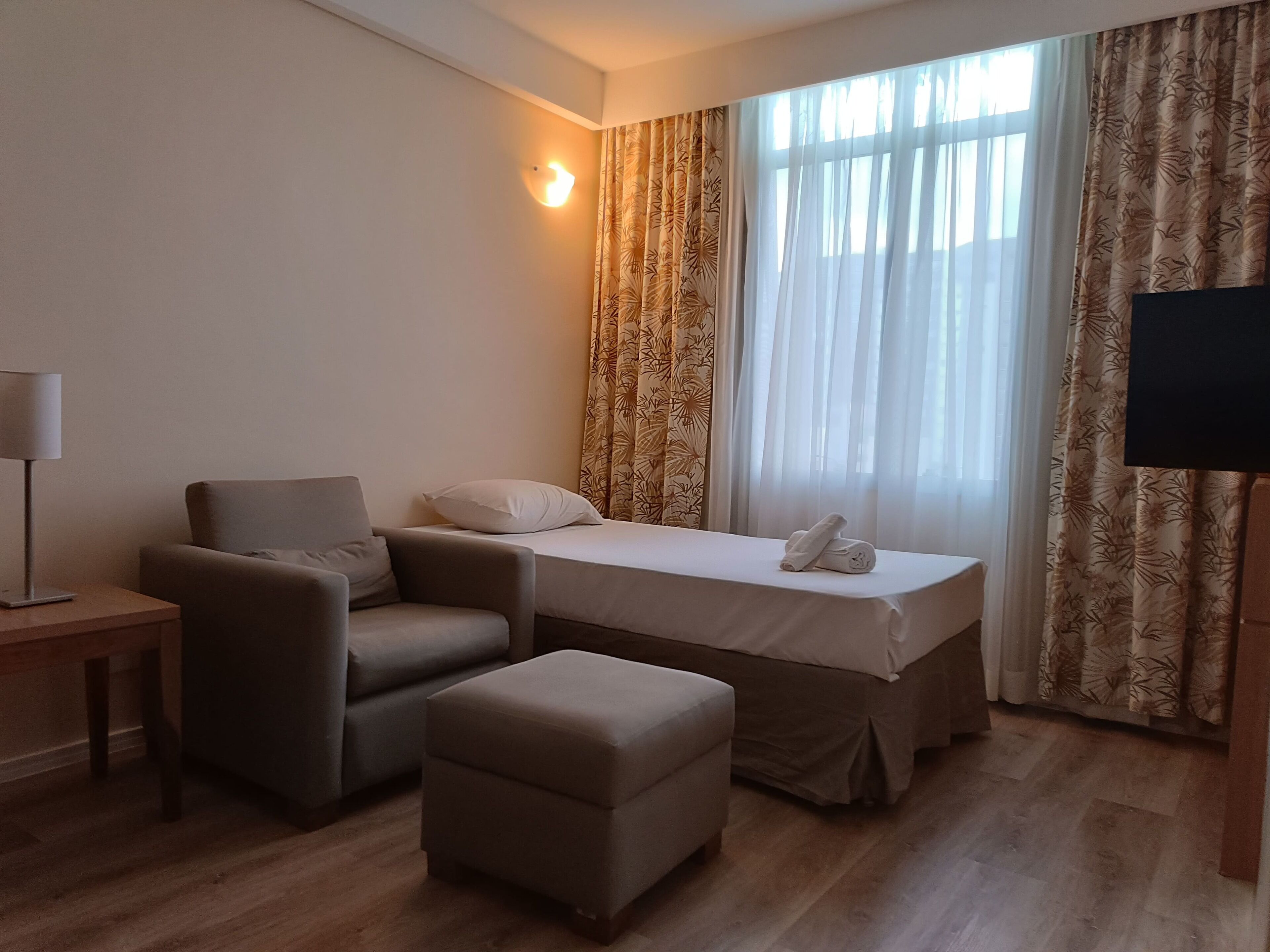 Quarto de hotel com cama, poltrona, banquinho, mesa de cabeceira, luminária e televisão.