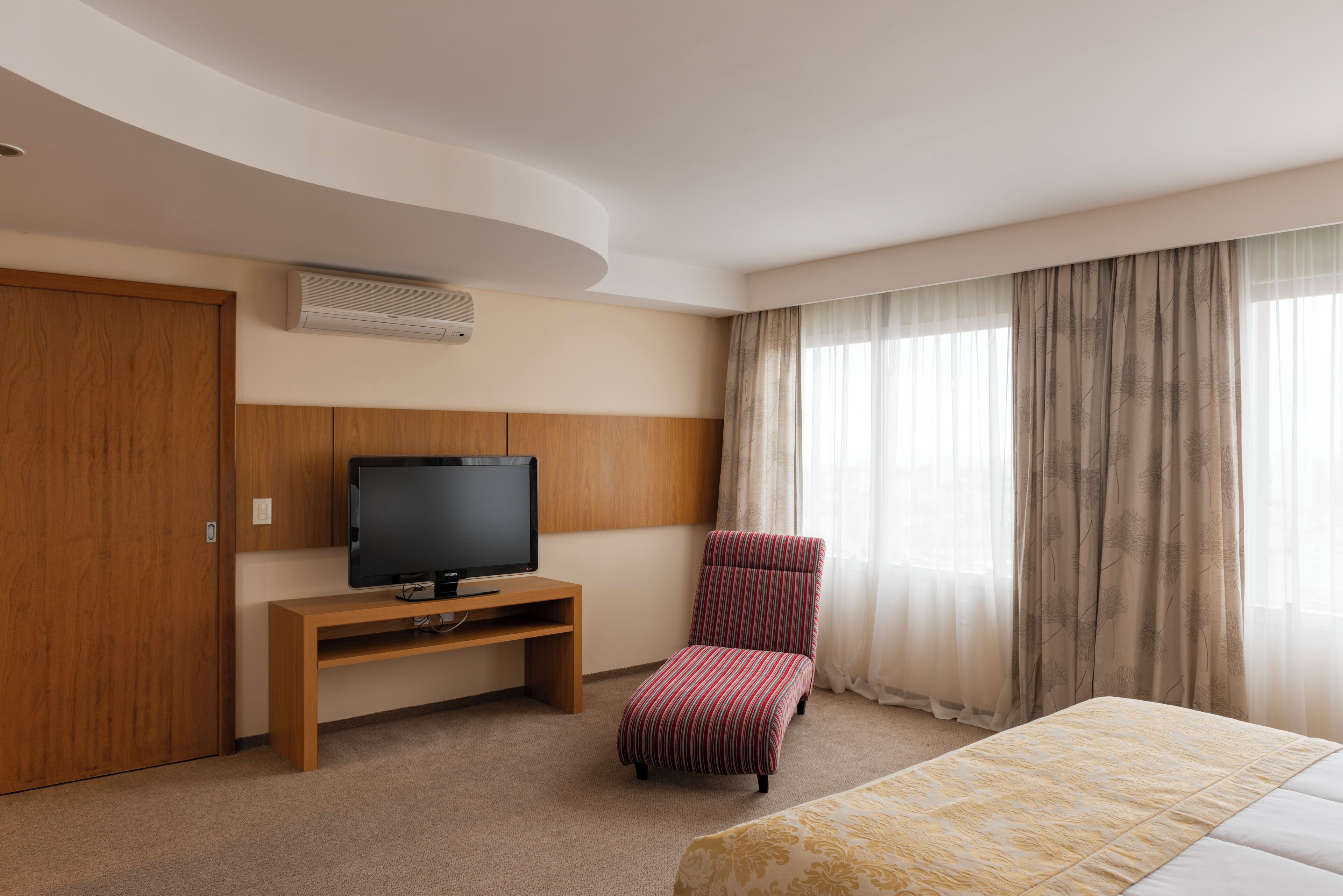 Ein Hotelzimmer mit einem Bett, einem Fernseher auf einem Holzgestell, einem rot gestreiften Sessel und einem großen Fenster mit transparenten Vorhängen.