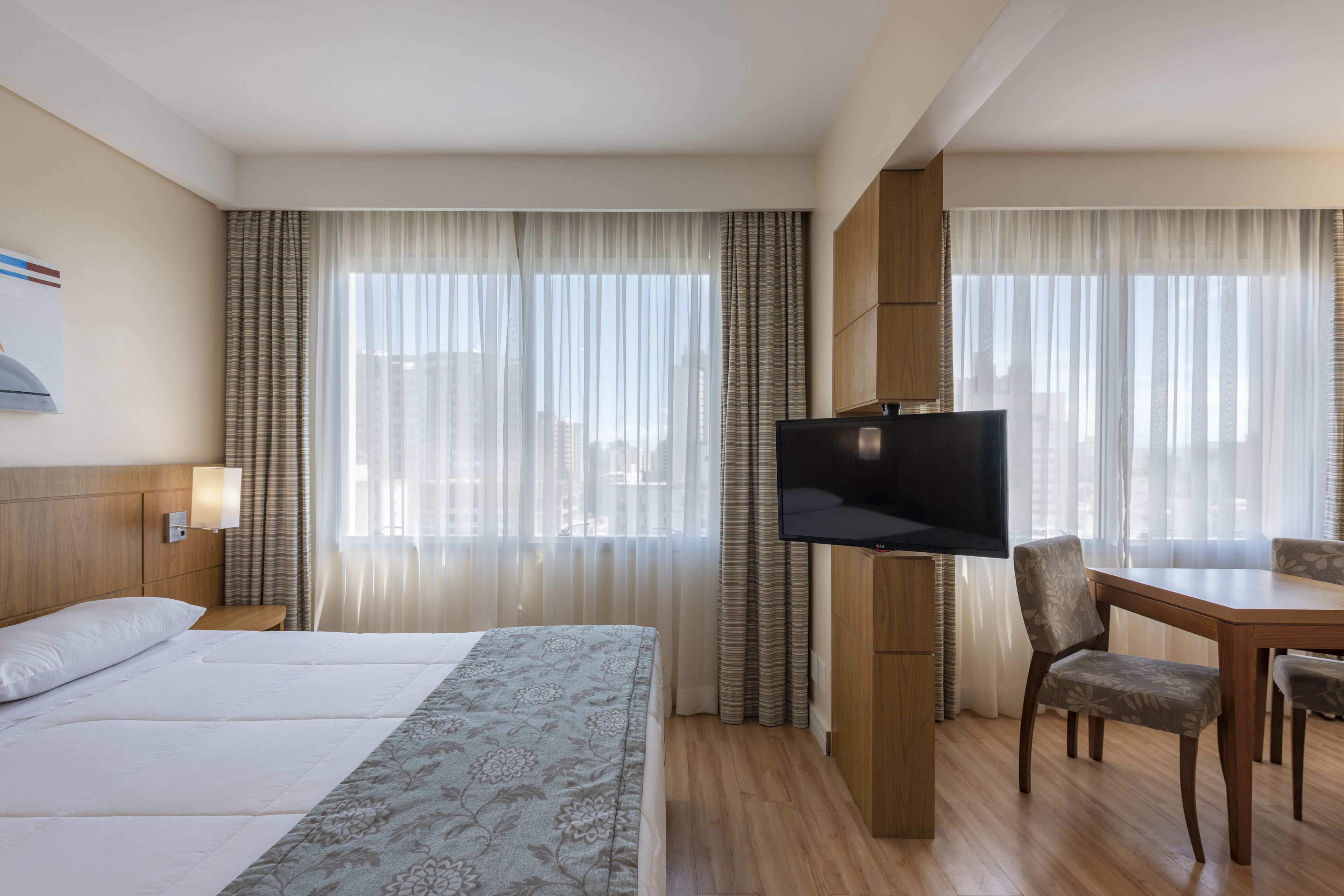 A hotel room with a large bed, a TV, a dining table with chairs, and a view of the city through sheer curtains.