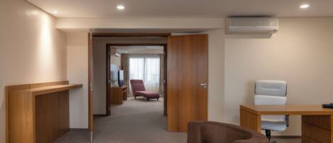 Une chambre d’hôtel avec un bureau en bois, une chaise marron, un fauteuil de bureau blanc, une télévision et un appareil de climatisation.