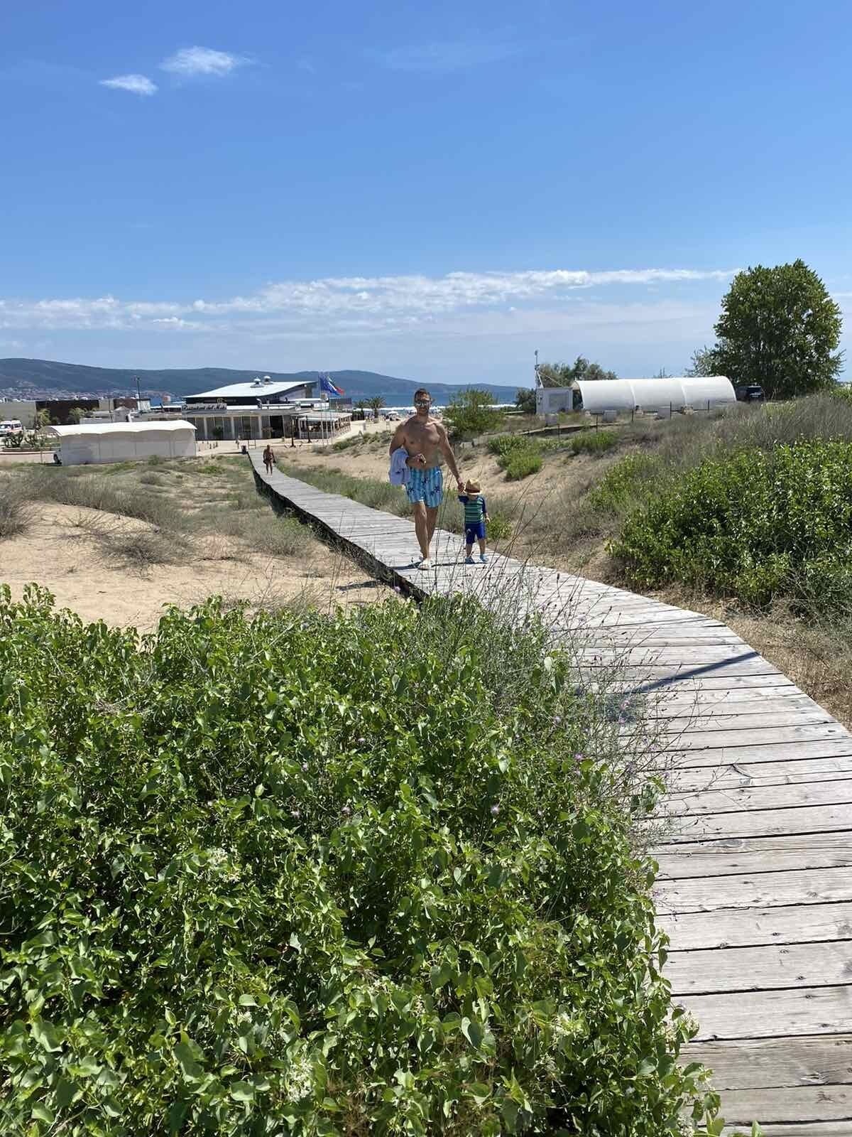 A wooden boardwalk leading to a beach area with a person and a child walking.
