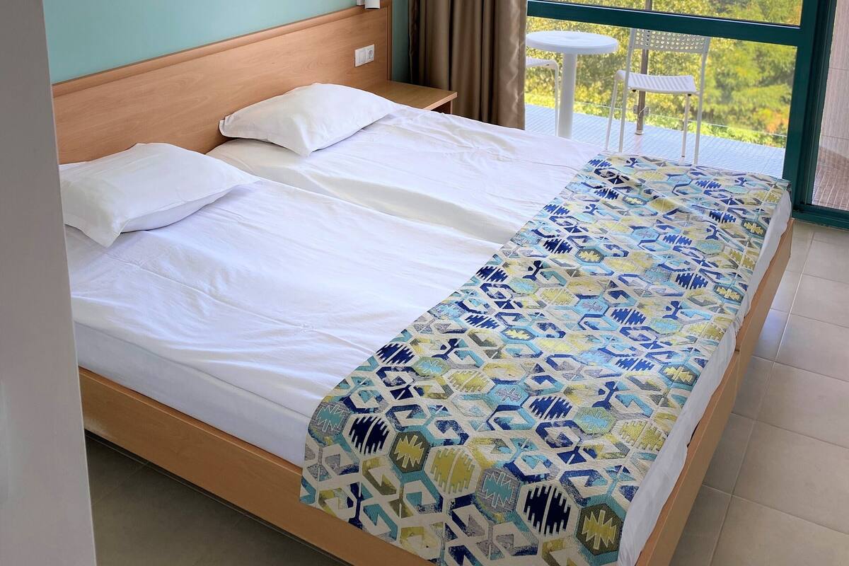 Une chambre d’hôtel avec un lit, une fenêtre avec des rideaux, un appareil de climatisation et un balcon avec une table et une chaise.