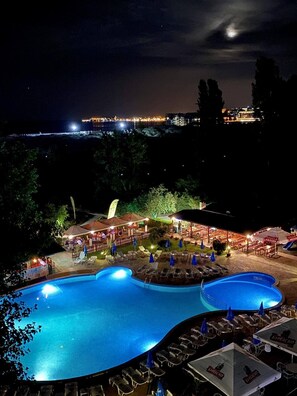 A nighttime view of a resort with a large swimming pool, outdoor seating areas, and a view of the city skyline. - Sirena Hotel (Sunny Beach)