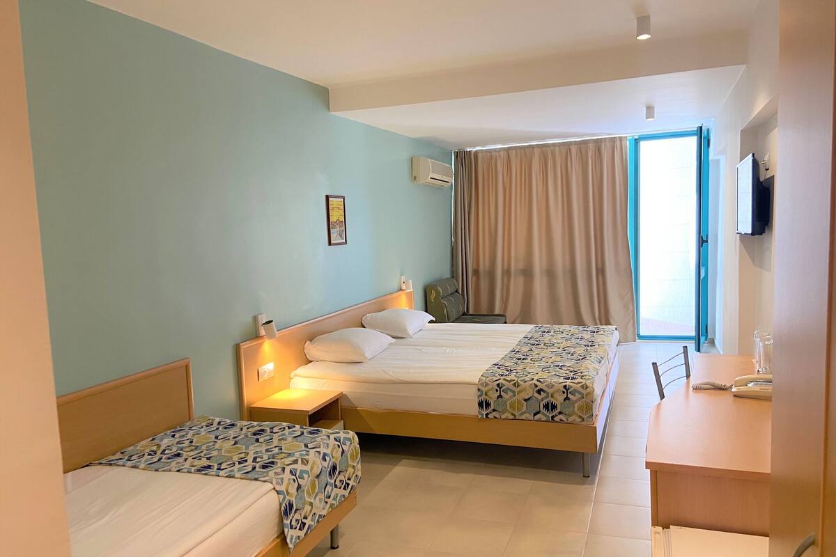 Une chambre d’hôtel avec deux lits, un bureau et une chaise. Il y a une fenêtre avec des rideaux, un climatiseur et un téléviseur fixé au mur.