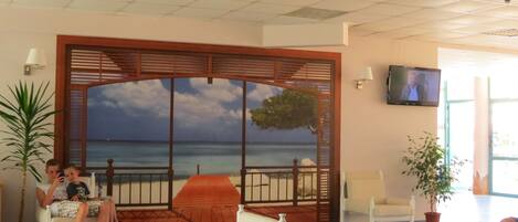 A room with a view of the sea through a large window, a wooden partition, a television on the wall, and a glass table.