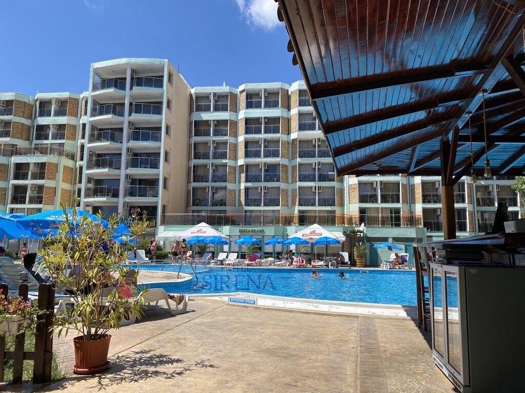 A hotel pool area with a large pool, umbrellas, and a building with multiple balconies in the background.