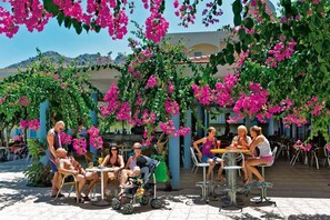 Un área de estar al aire libre con mesas y sillas, rodeada de follaje y flores rosadas.
