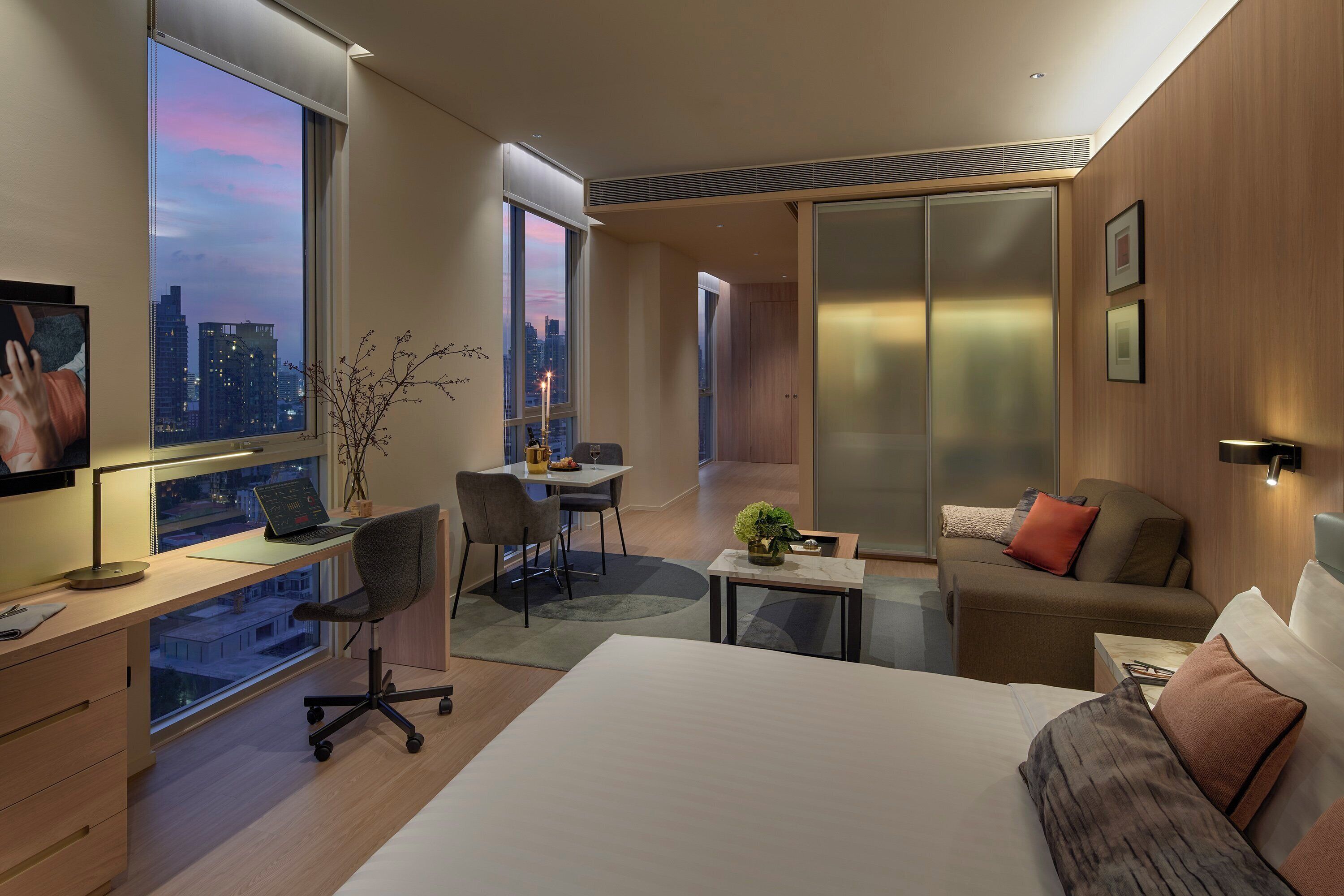Habitación de hotel moderna con una cama grande, un escritorio con computadora, un área de comedor y vistas a la ciudad.