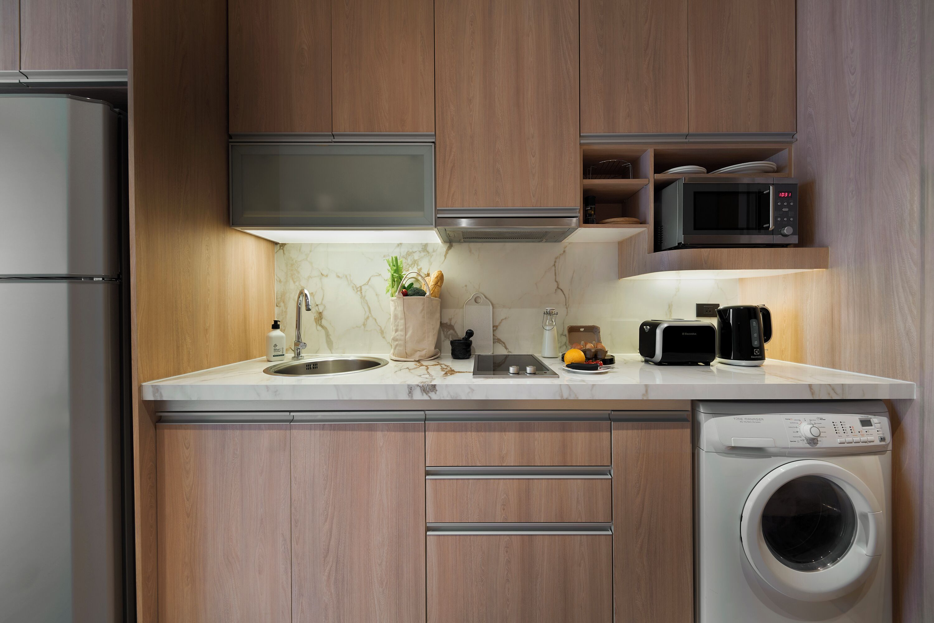 Una cocina moderna con armarios de madera, un refrigerador de acero inoxidable, un microondas, un lavamanos y una lavadora integrada.