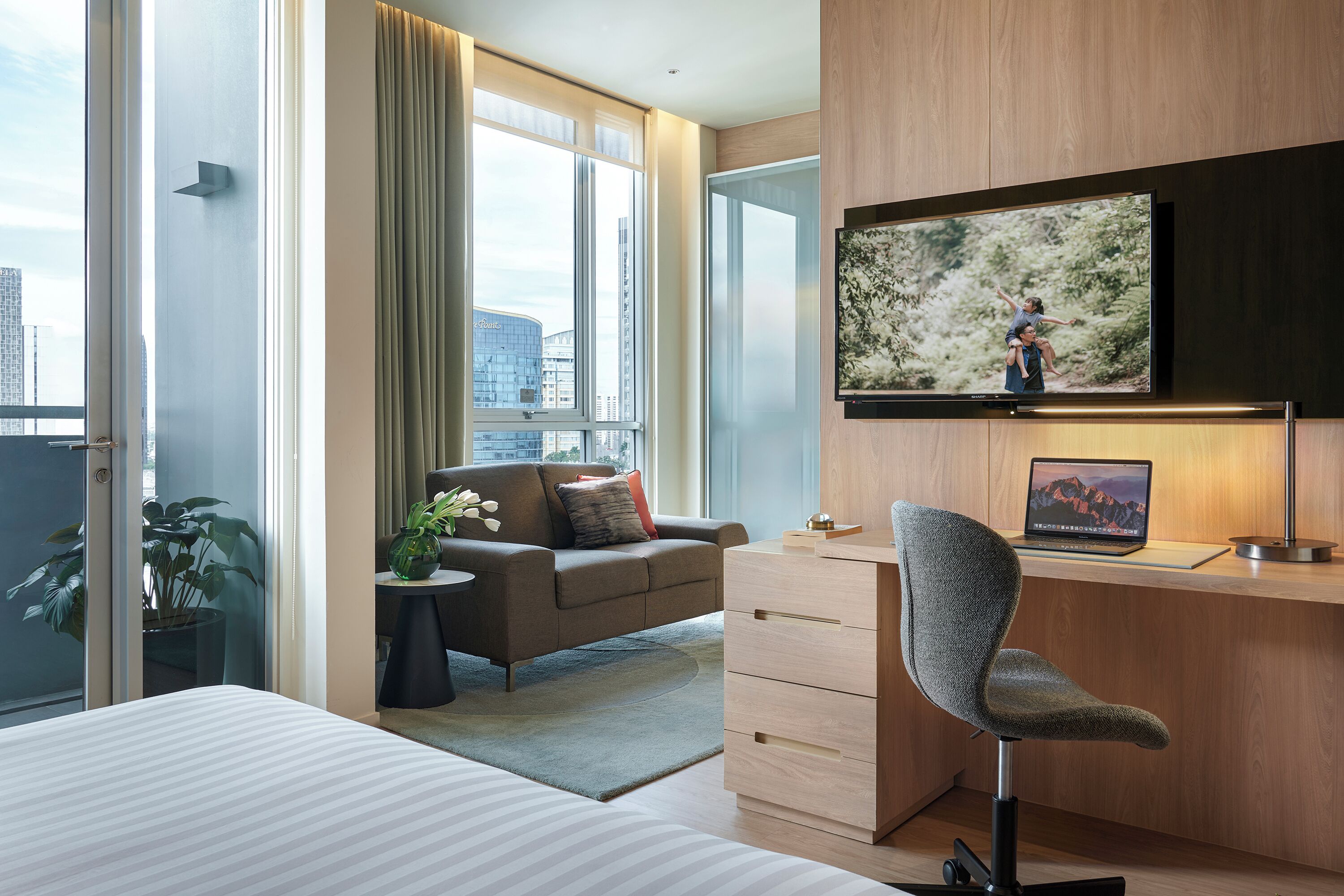 A modern hotel room with a large bed, a desk with a laptop, a TV on the wall, and a view of the city through large windows.