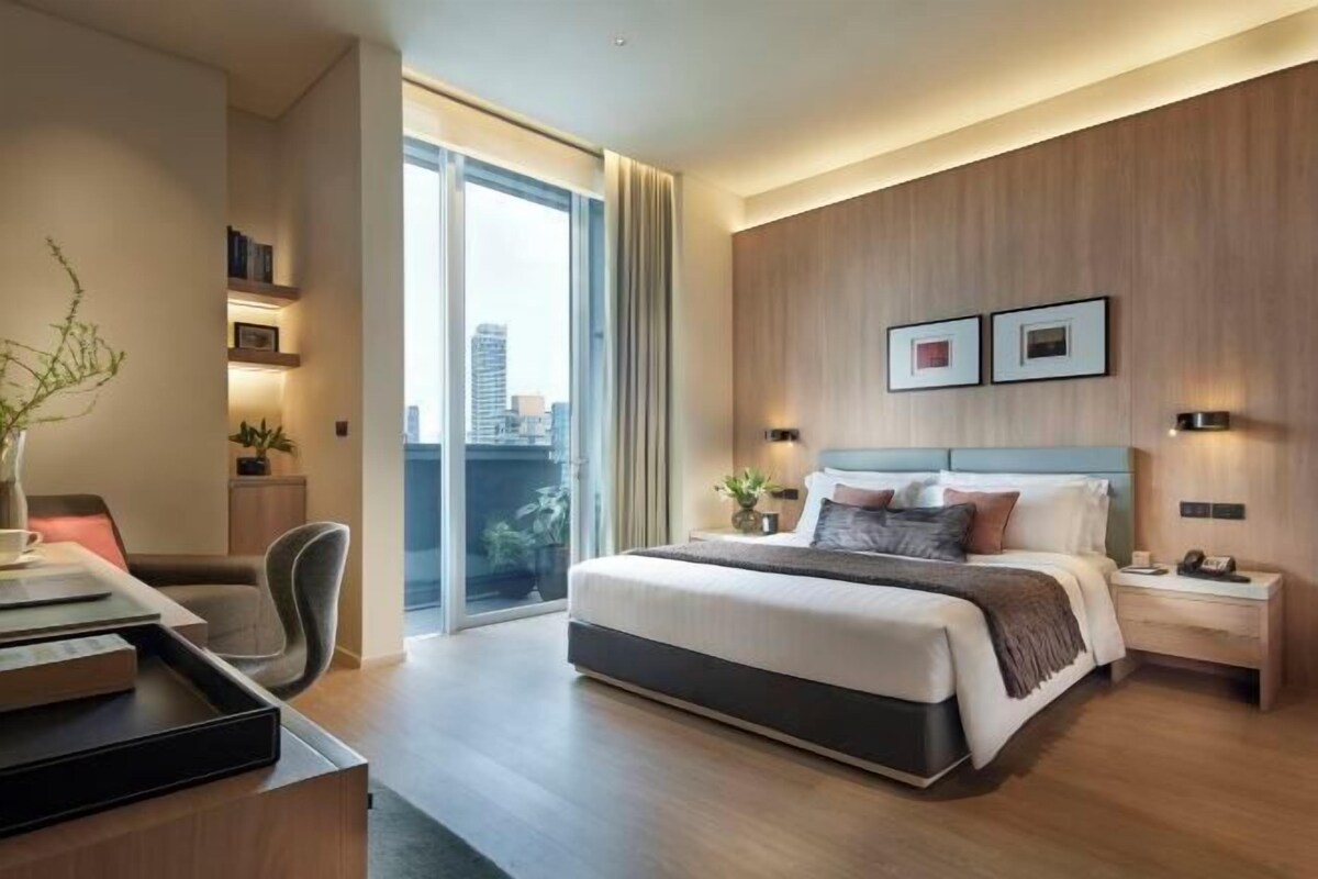 A modern hotel room with a large bed, a desk with a chair, a view of the city through a large window, and a wooden wall panel.
