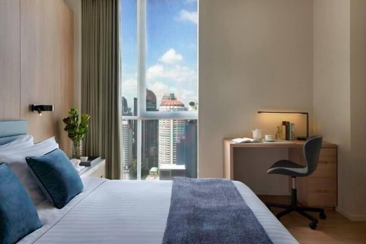 A hotel room with a bed, a desk, a chair, a window with a city view, and a plant.